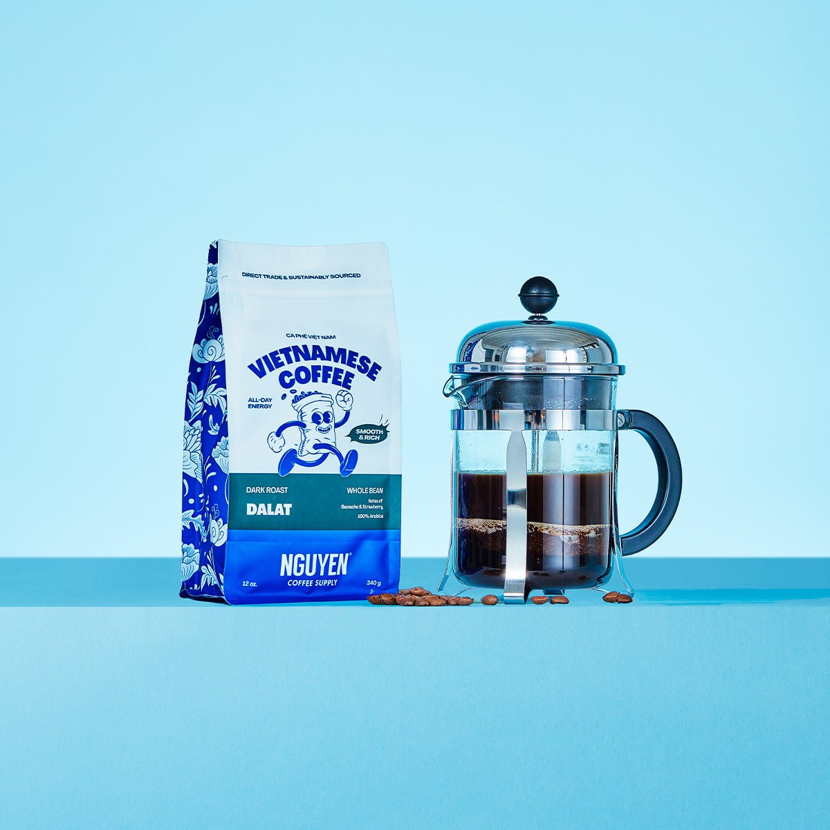 A bag of Nguyen Vietnamese Coffee Dalat whole bean coffee stands next to a glass French press filled with brewed coffee on a light blue background.