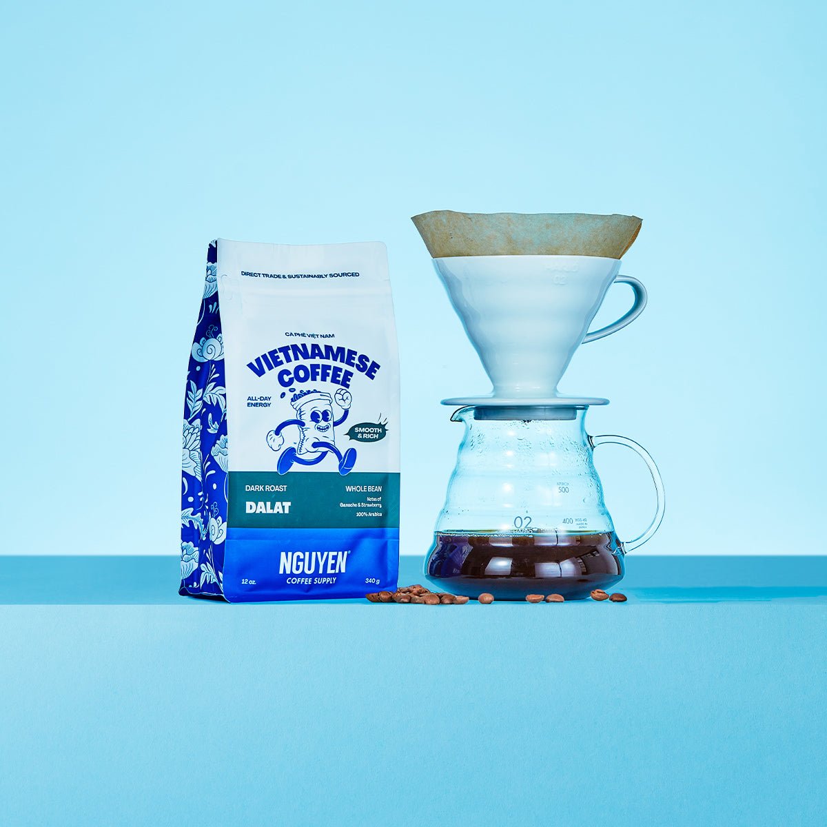 A bag of Nguyen Vietnamese Coffee Dalat whole bean coffee stands next to a pour-over setup with a white cone filter, glass carafe, and scattered coffee beans on a light blue background.