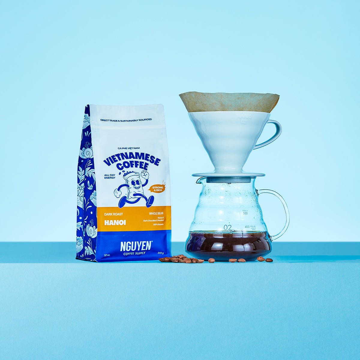 A bag of Nguyen Vietnamese Coffee Hanoi dark roast coffee stands next to a white pour-over dripper and glass carafe filled with brewed coffee, with a few coffee beans scattered on a light blue surface and background.