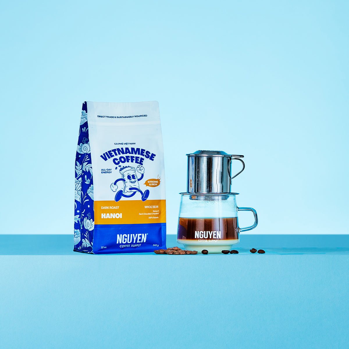 A bag of Nguyen Vietnamese Coffee Hanoi dark roast whole bean coffee stands next to a traditional phin filter coffee maker over a glass of brewed coffee with sweetened condensed milk, with a few coffee beans scattered in between, on a light blue background.