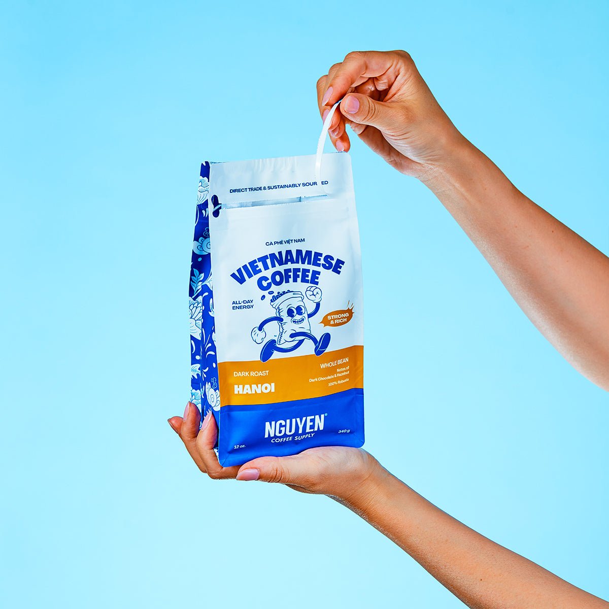 A person holds a bag of Nguyen Vietnamese Coffee Hanoi against a solid light blue background, with one hand holding the bag and the other pulling the white drawstring.