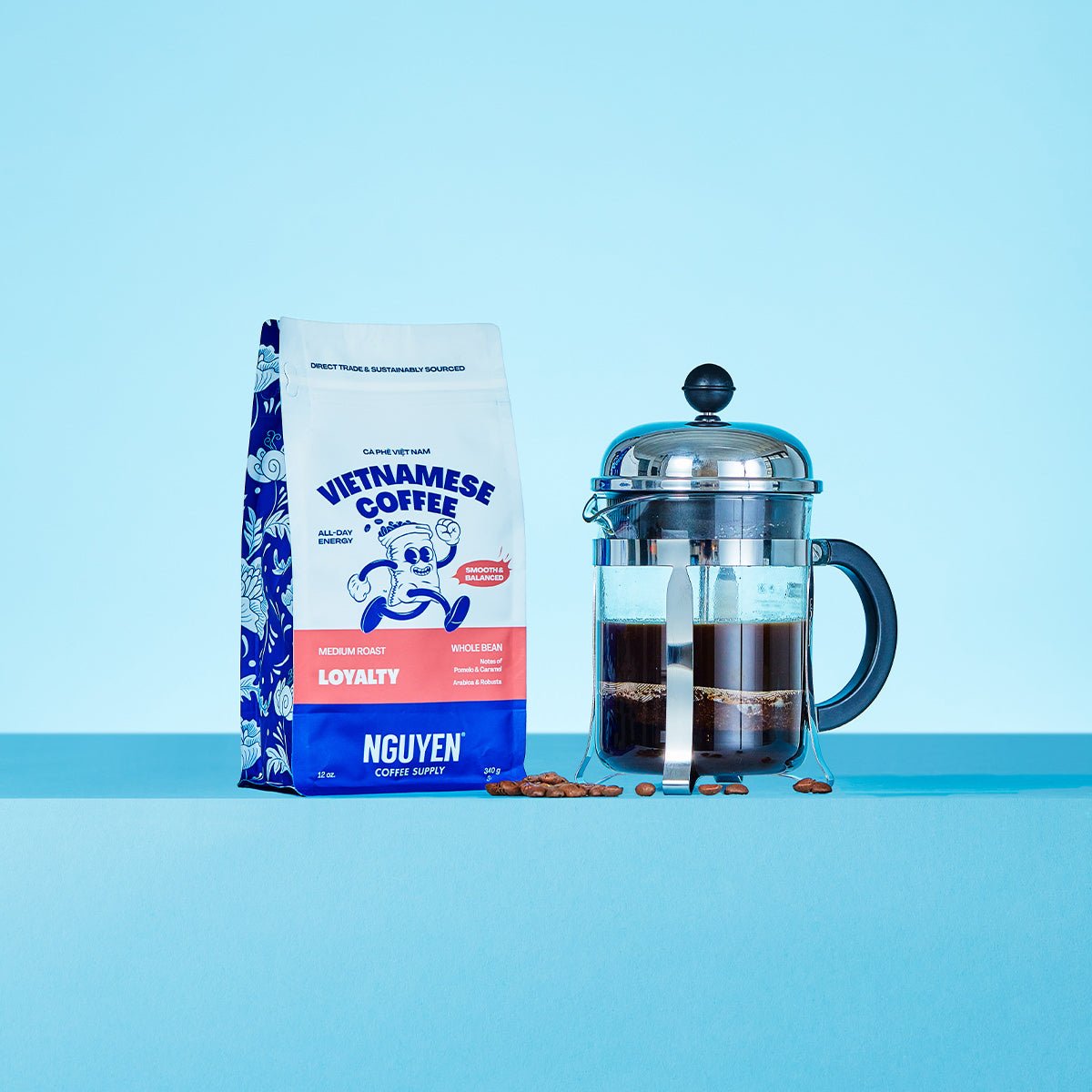 A bag of Nguyen Vietnamese Coffee Loyalty medium roast coffee beans stands next to a glass French press filled with dark coffee on a light blue background.