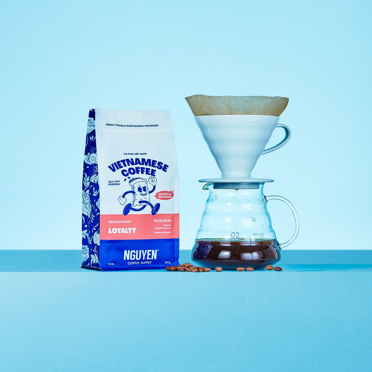 A bag of Nguyen Vietnamese Coffee Loyalty medium roast coffee beans stands next to a white pour-over dripper and glass carafe filled with brewed coffee, with a few coffee beans scattered in between, set against a light blue background.