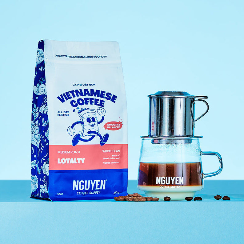 A bag of Nguyen Vietnamese Coffee Loyalty medium roast coffee beans stands next to a traditional phin filter dripping on top of a glass filled with brewed coffee and sweetened condensed milk, a few coffee beans scattered in between, on a light blue background.