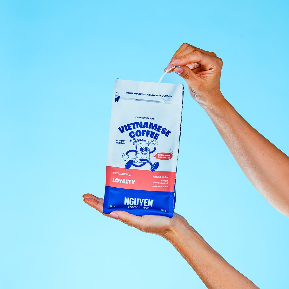 A person holds a bag of Nguyen Vietnamese Coffee Loyalty medium roast coffee against a solid light blue background, pulling the tab to open the bag