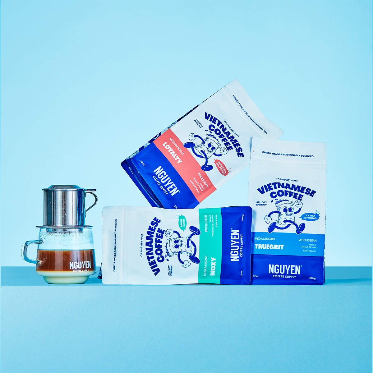Four bags of Nguyen Vietnamese Coffee beans in various flavors and roasts, arranged artistically next to a phin filter glass pour-over coffee maker on a light blue background.