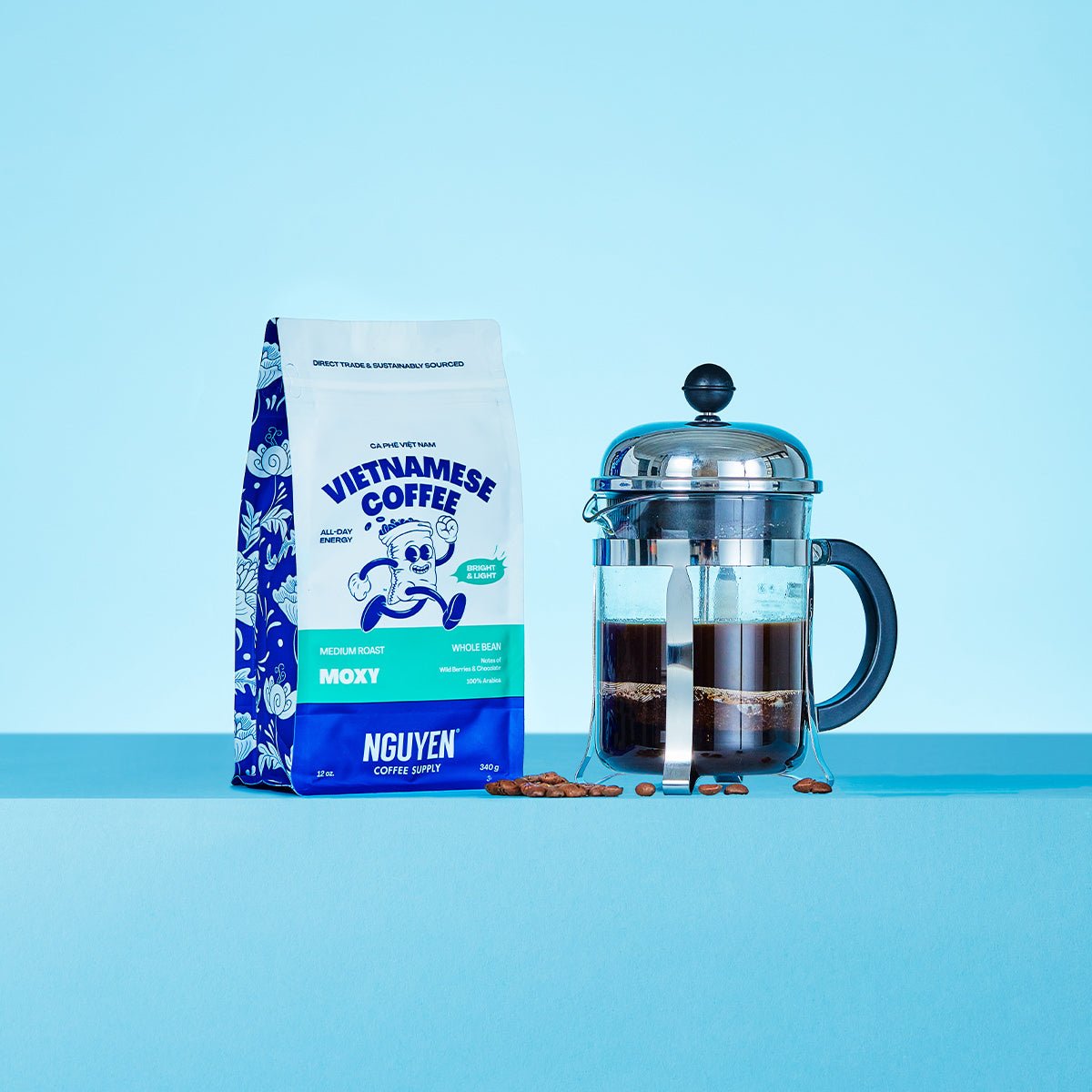 A bag of Nguyen Vietnamese Coffee Moxy medium roast whole bean coffee stands next to a glass French press filled with dark coffee, with a few coffee beans scattered in between, set against a solid light blue background.