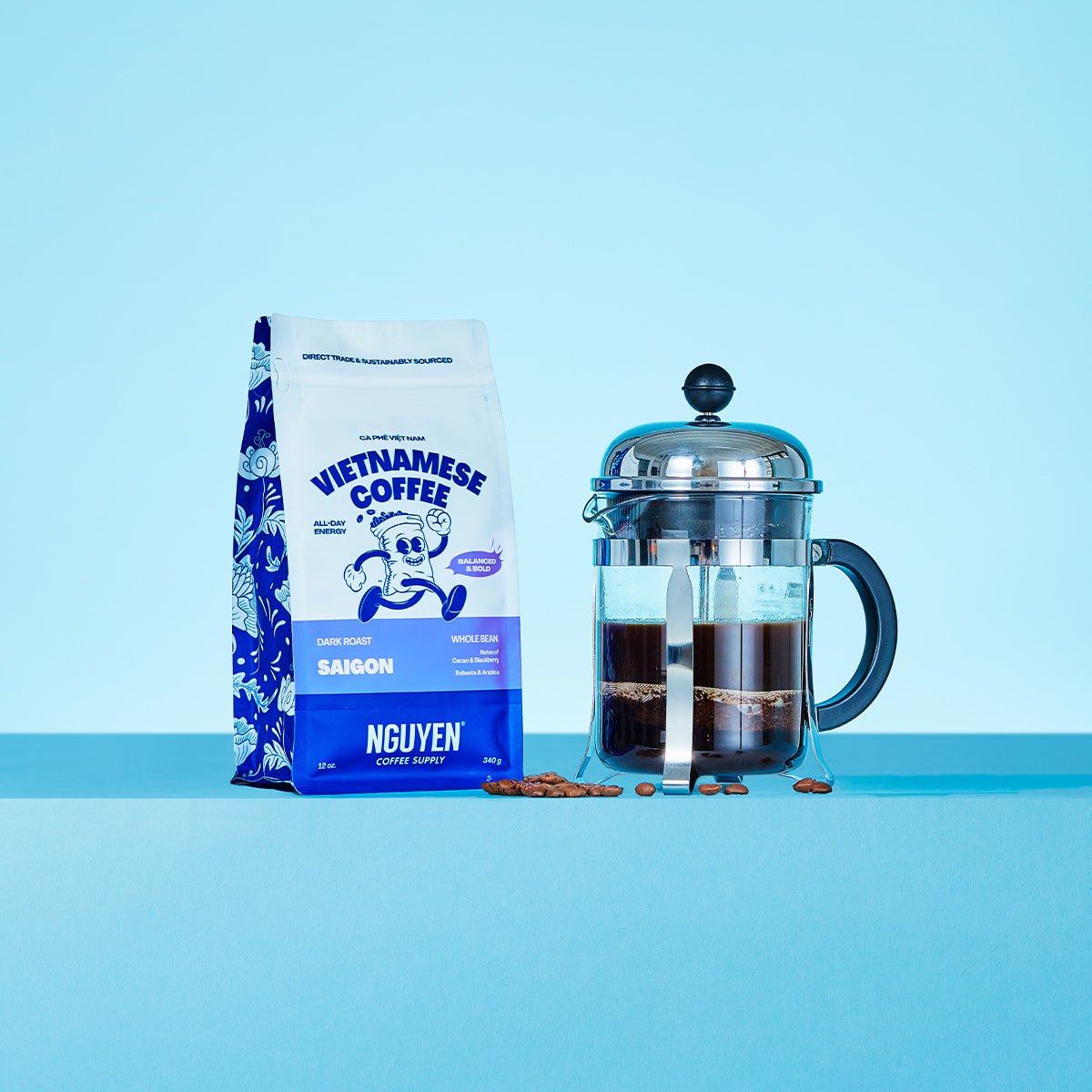 A bag of Nguyen Vietnamese Coffee Saigon whole bean dark roast coffee stands next to a glass French press filled with brewed coffee on a light blue background.