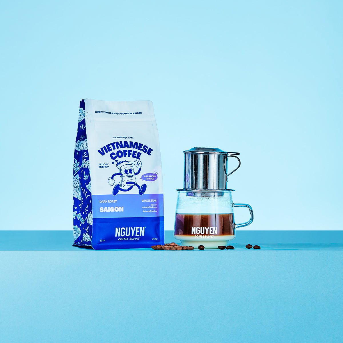 A bag of Nguyen Vietnamese Coffee Saigon dark roast next to a traditional phin filter dripping over a glass with brewed coffee and sweetened condensed milk, with a few coffee beans scattered on a light blue background.