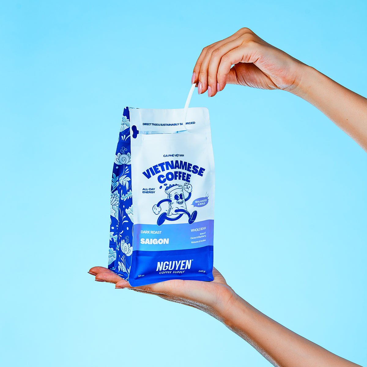 A hand holds a blue and white bag of Nguyen Vietnamese Coffee Saigon dark roast while another hand pulls the tab to open the bag, set against a solid light blue background.