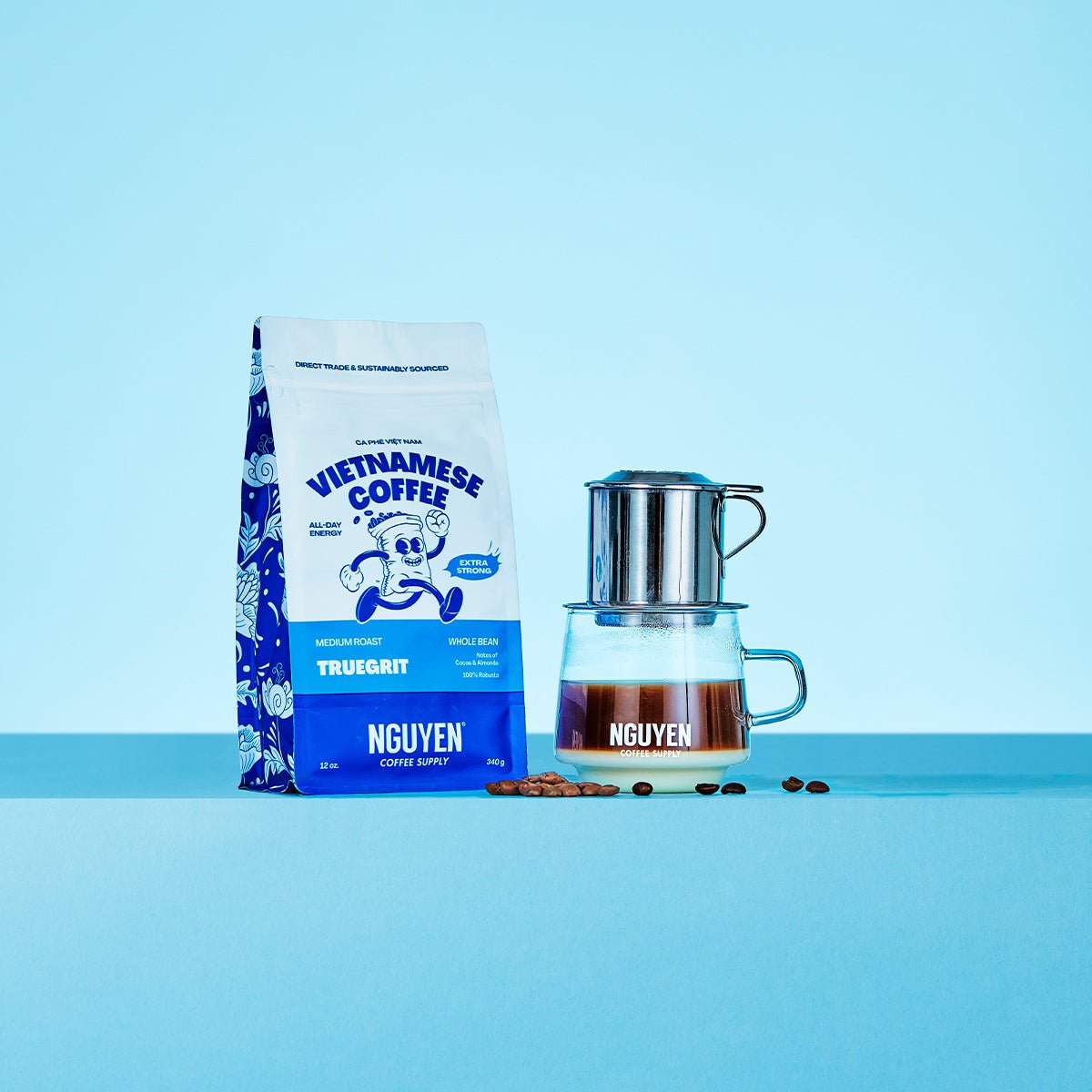 A bag of Nguyen Vietnamese coffee Truegrit medium roast extra strong next to a traditional phin filter on top of a glass cup filled with brewed coffee and sweetened condensed milk, with coffee beans around
