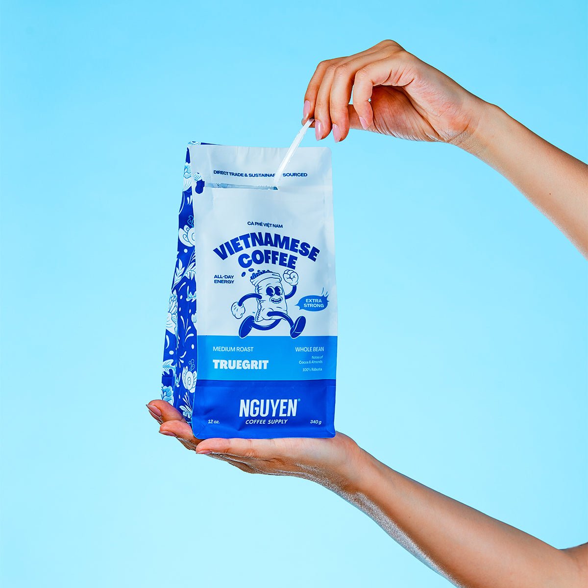 A person holds a bag of Nguyen Vietnamese Coffee TrueGrit Medium Roast Single whole coffee against a light blue background, with one hand holding the bag and the other pulling the tab open